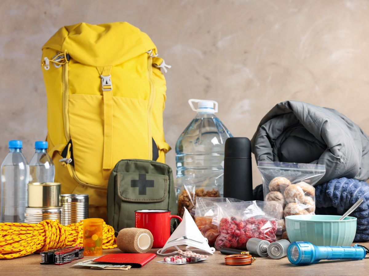 Emergency preparedness supplies including a yellow backpack, canned food, water, flashlight, and first aid kit — representing disaster preparedness for seniors.