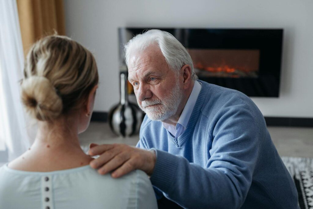An elderly man comforts a woman indoors near a cozy fireplace, expressing emotional support.