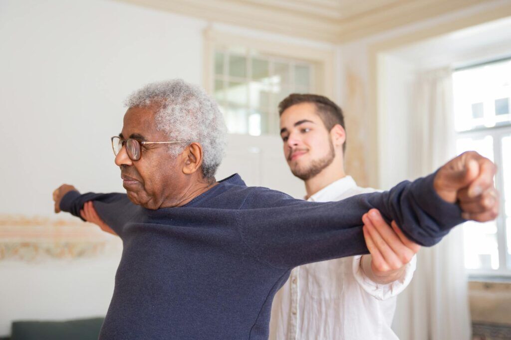 Senior man receiving caregiver support for stretching, strength, and rehabilitation indoors.